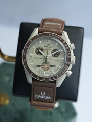 A Swatch x OMEGA MoonSwatch Collection watch with a brown leather band and a brown and silver watch face, displayed on a stand with the OMEGA logo.
