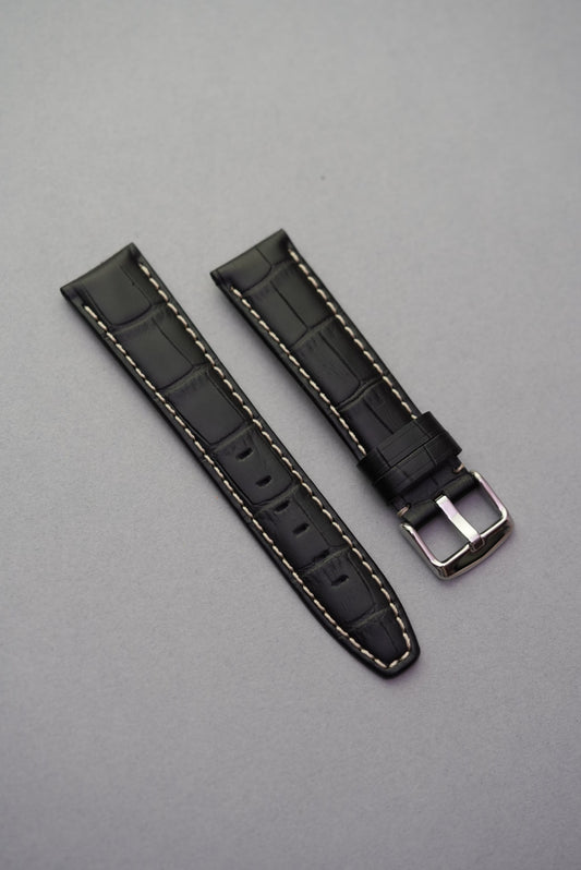 Black alligator-style leather watch strap with white contrast stitching and polished silver buckle, displayed on a grey background.