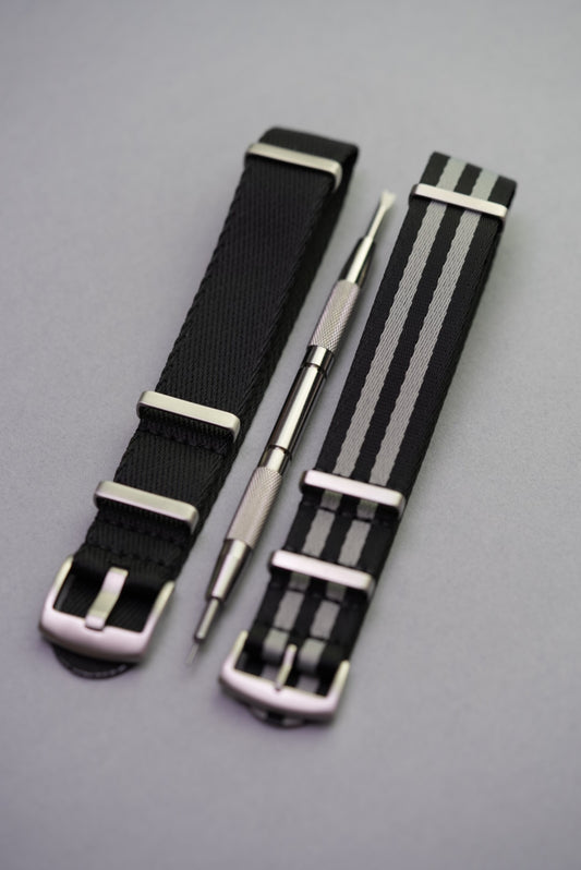 Top-down view of a black NATO strap, grey-striped NATO strap, and spring bar tool arranged diagonally on a grey background.