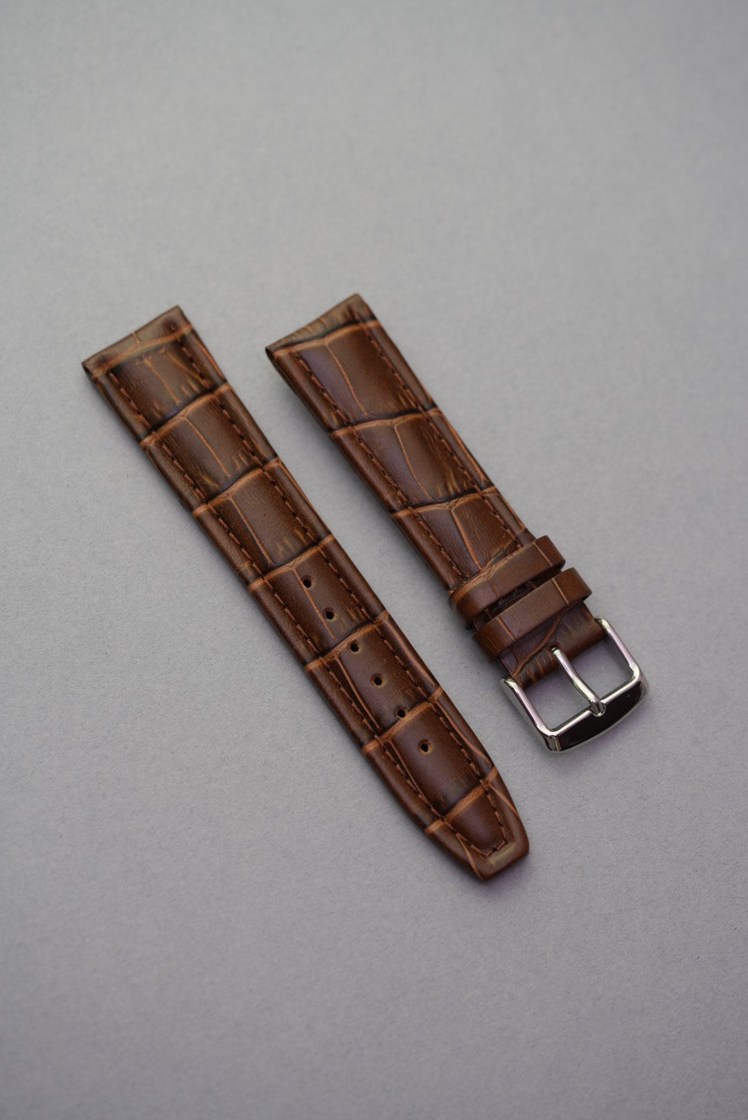 Brown alligator-style leather watch strap with visible stitching and silver buckle, presented on a grey background.