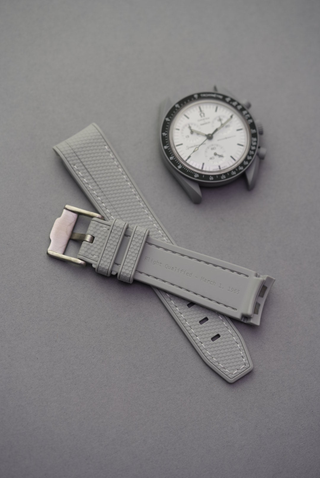 Grey Flight Qualified 1965 strap for MoonSwatch with white stitching and brushed steel buckle, displayed with MoonSwatch Mission to the Moon.