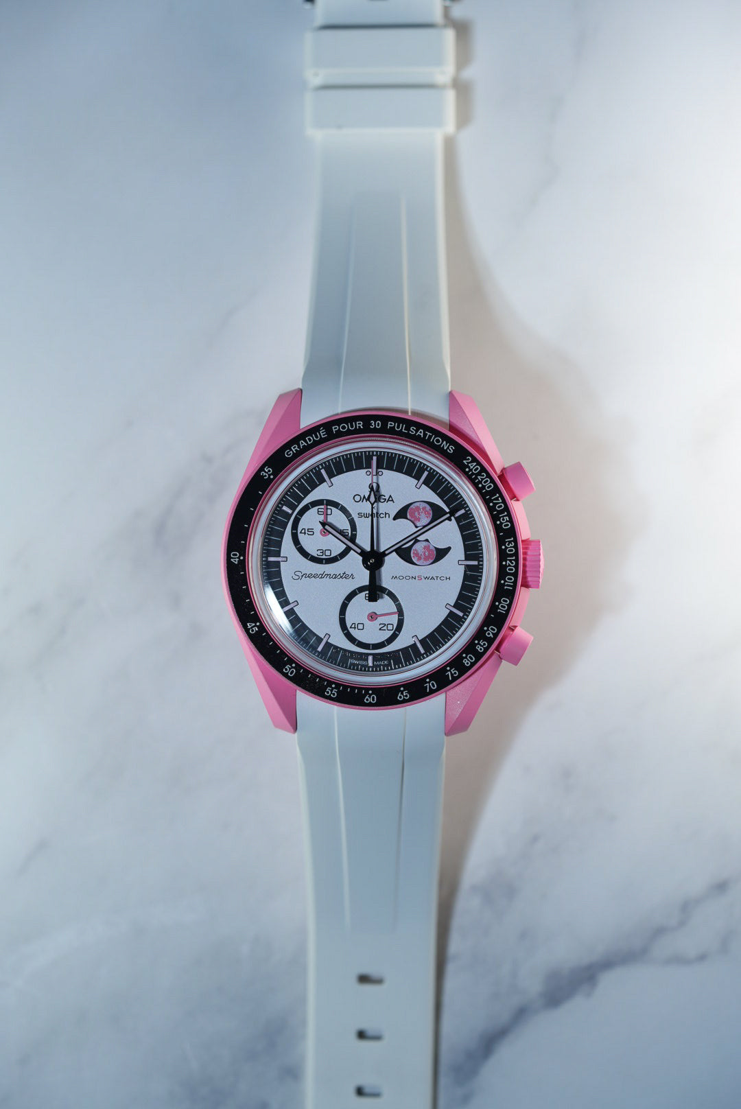 Top-down flat lay of the Swatch x Omega Mission to the Moonphase Pink with white rubber strap – set against a marble background.