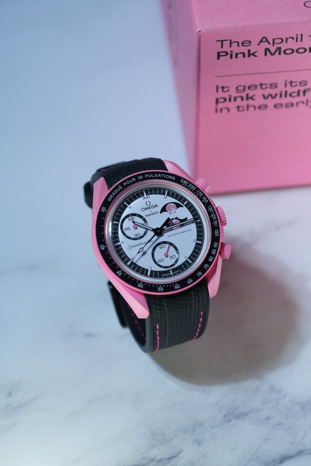 Lifestyle shot of the Pink Moonphase MoonSwatch placed in front of its original pink box – paired with a black strap and pink stitching.Lifestyle shot of the Pink Moonphase MoonSwatch placed in front of its original pink box – paired with a black strap and pink stitching.