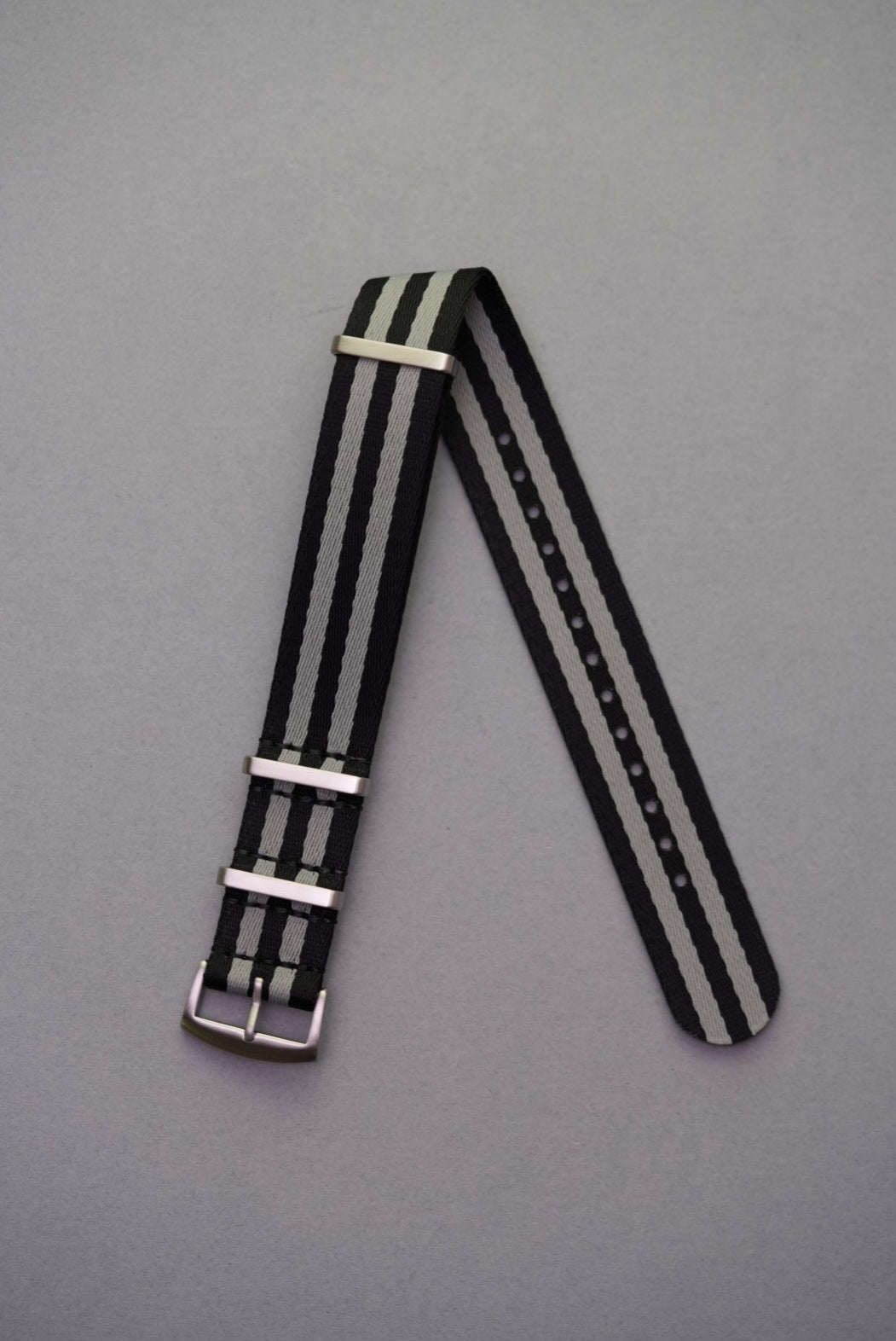 Premium black and grey NATO watch strap in Bond-style design with brushed metal keepers, laid flat on grey background. Fabric strap replacement for Swatch x OMEGA Mission To The Moon MoonSwatch