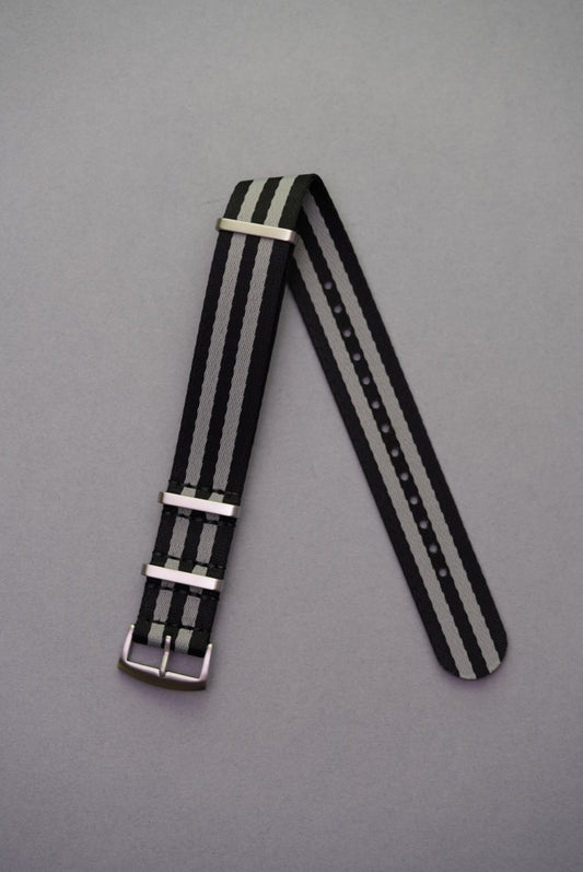 Premium black and grey NATO watch strap in Bond-style design with brushed metal keepers, laid flat on grey background. Fabric strap replacement for Swatch x OMEGA Mission To The Moon MoonSwatch