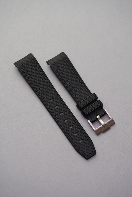 A black curved rubber watch strap with a stainless steel buckle laid out flat on a grey background.