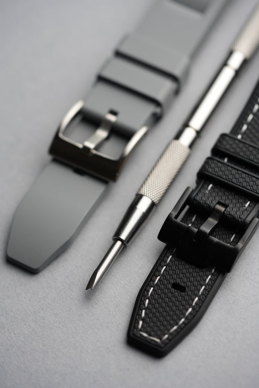 Detailed view of strap buckles on a grey rubber strap and black textured strap, with a precision strap-changing tool in the middle.