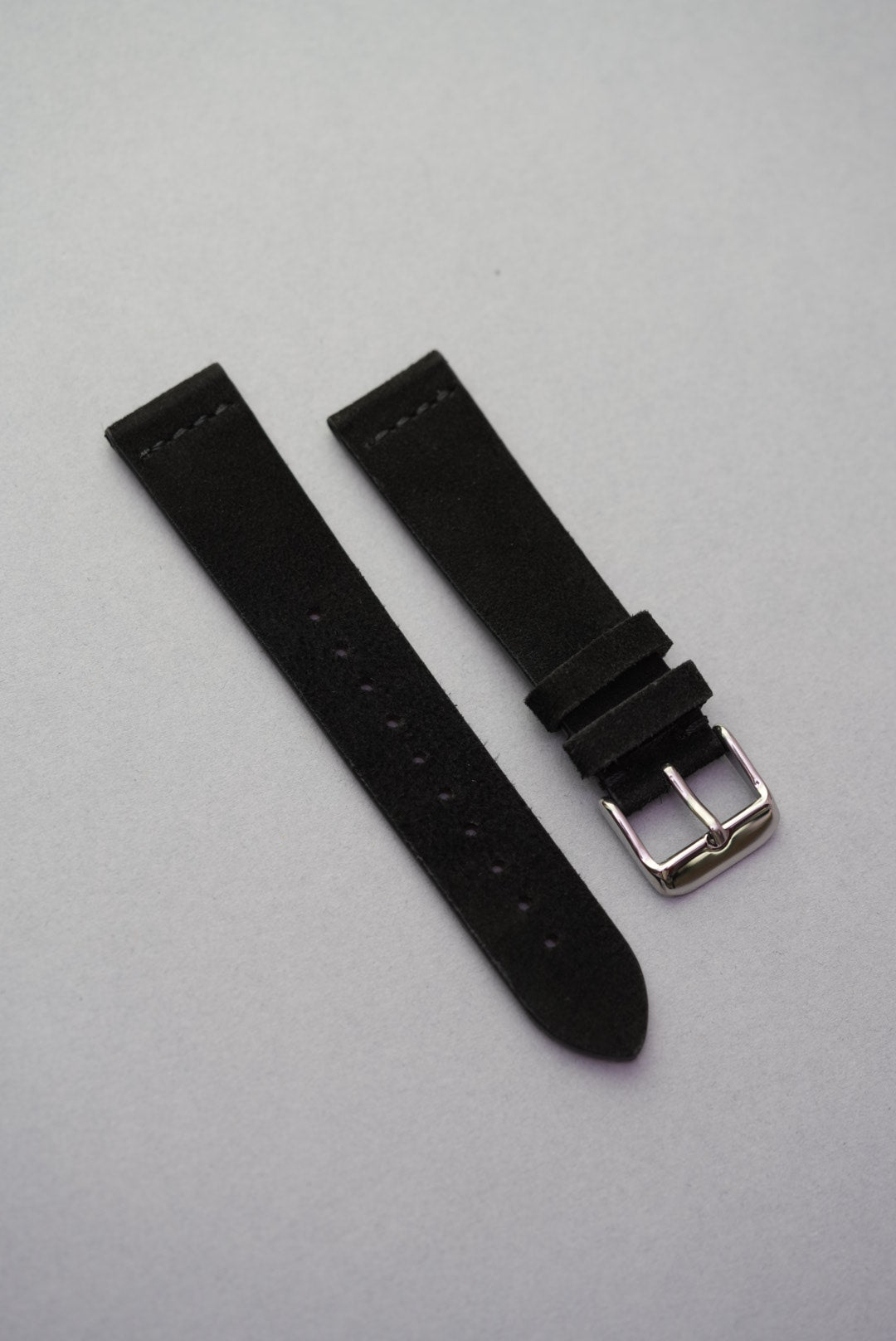 Black suede leather watch strap with subtle blue stitching and polished silver buckle, displayed flat on a light grey background.