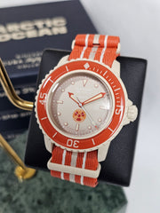 A close-up photograph of the Blancpain x Swatch Scuba Fifty Fathoms "Arctic Ocean" watch displayed on a black stand. The watch features a beige Bioceramic case, an orange bezel with white markings, and a textured silver dial with luminescent hands and hour markers. A distinct red and yellow "No Radiations" logo is positioned above 6 o’clock. It is paired with an orange NATO-style strap with white and beige stripes. In the background, a blurred box with the words "Arctic Ocean" reinforces the model's theme.