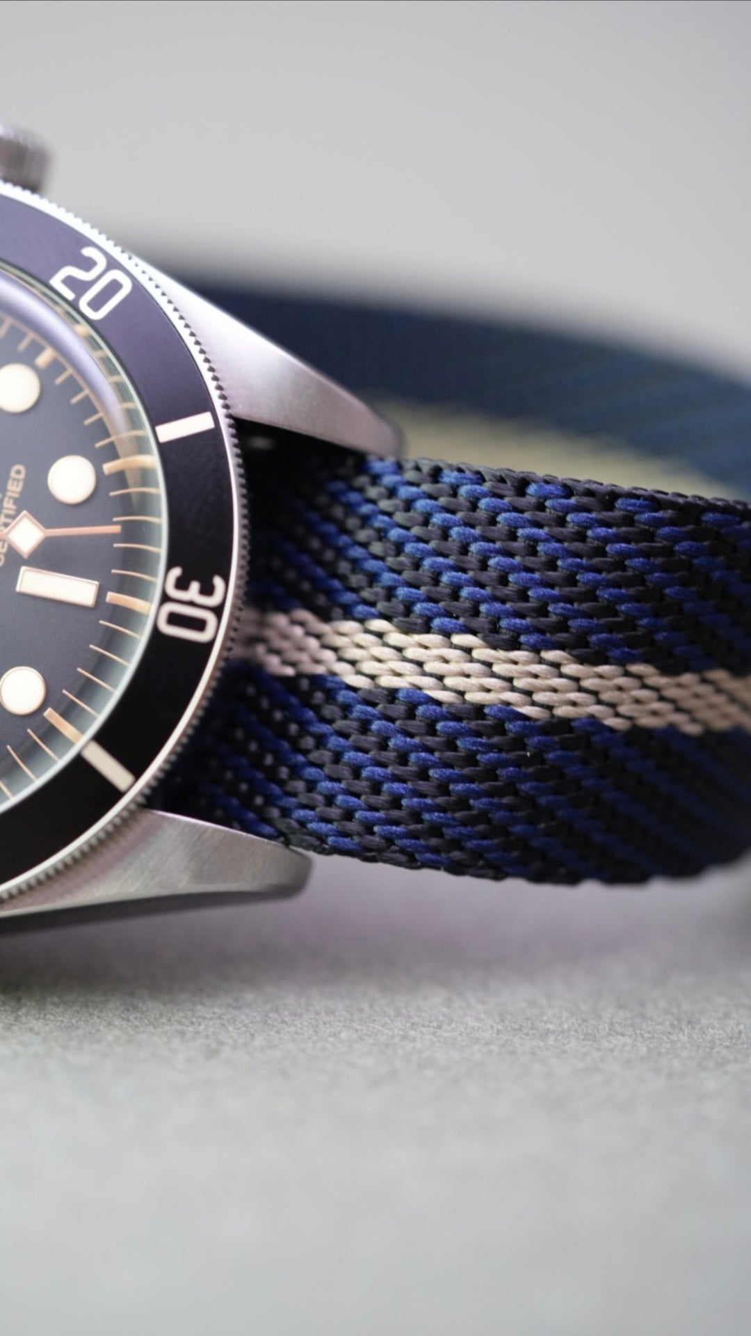 Tudor Black Bay laid flat with a black, blue, and cream woven nylon strap on a grey background.