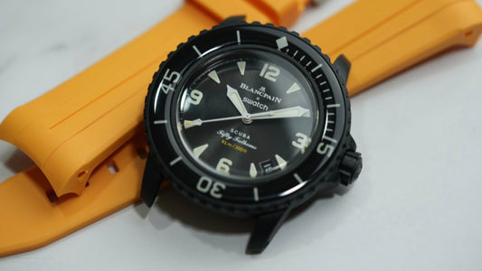 Blancpain x Swatch Scuba Fifty Fathoms watch with black dial and bezel, placed on an orange rubber strap. The watch features bold numerals, luminescent hands, and a rotating dive bezel, with "91m / 300ft" water resistance marking on the dial.