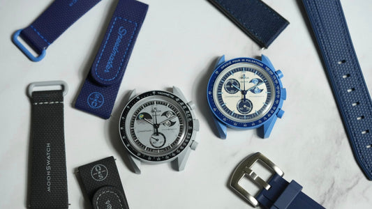 An image of two MoonSwatch watch heads, surround by different types of straps most in black an blue colours on a marble white background