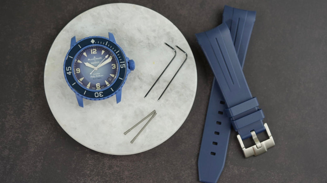 A blue Blancpain x Swatch Scuba Fifty Fathoms watch case displayed on a white circular surface, accompanied by a set of watch tools and a matching blue rubber watch strap with a silver buckle, all set against a dark textured background