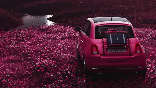 Pink Fiat with Swatch x Omega branding parked in a surreal pink landscape, promoting the "Mission to the Pink Moon Phase" MoonSwatch release. Features #MoonSwatch text, highlighting the upcoming Swatch x Omega Pink Moon edition.