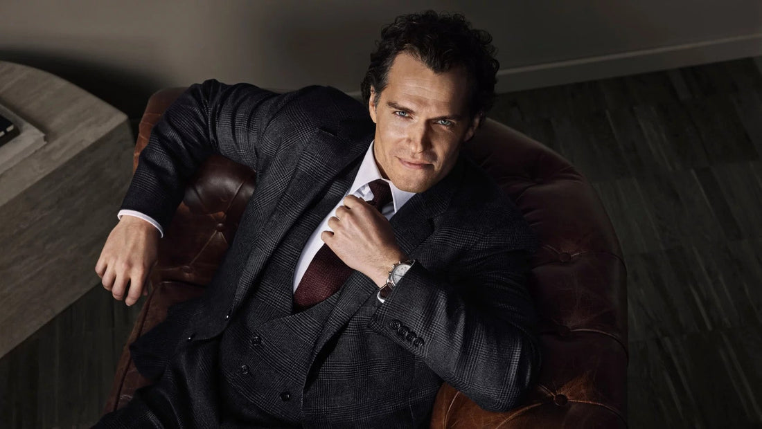 Henry Cavill wearing a Longines watch, dressed in a sleek black shirt, sitting in a dimly lit, elegant setting with a golden lamp in the background. His thoughtful expression adds to the refined atmosphere.