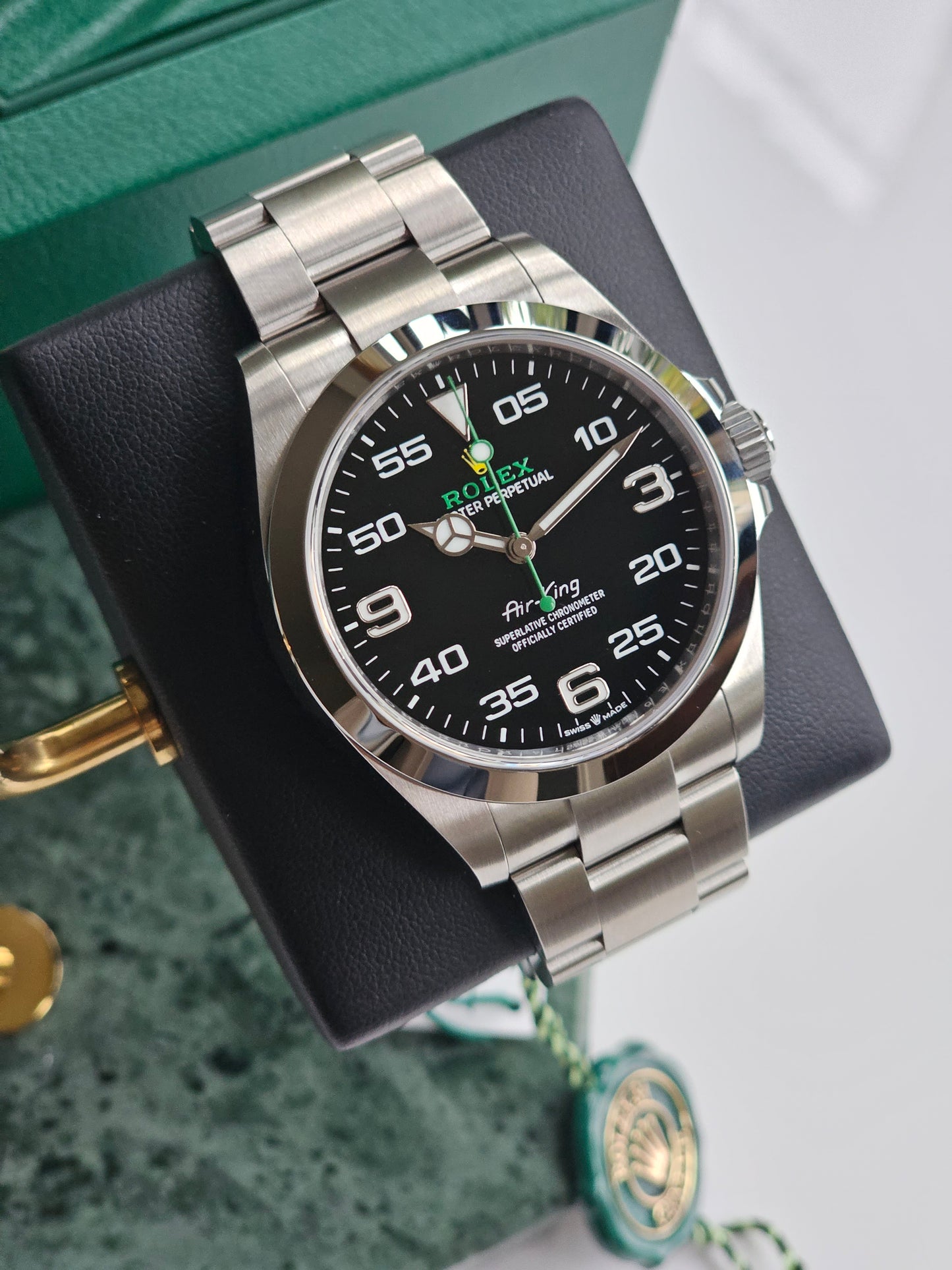 Rolex AirKing watch 126900, on a watch stand, time showing ten past ten. Watch angled to the left. Part of the green rolex box showing in the background