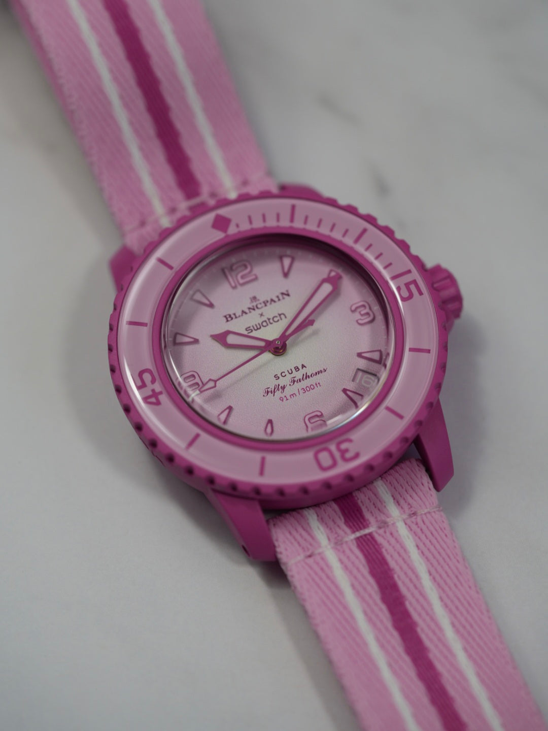 A Blancpain x Swatch Fifty Fathoms Scuba watch in vibrant pink, featuring a pink bioceramic case, a rotating bezel, a gradient pink dial with embossed numerals and indices, and a matching pink NATO-style strap with white stripes. The watch is water-resistant to 91m (300ft) and combines Swatch’s playful design with Blancpain’s iconic dive watch heritage.