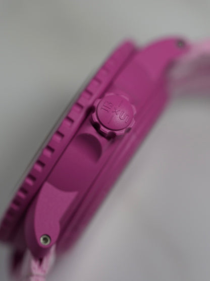 A side view of the Blancpain x Swatch Fifty Fathoms Scuba watch in pink crown, displayed on a black stand. The watch features a pink bioceramic case, a rotating bezel with dive markings, a gradient pink dial with embossed numerals, and a pink NATO-style strap with white stripes. The dial prominently displays the "Blancpain x Swatch" branding and "Fifty Fathoms Scuba" text, with a water resistance rating of 91m (300ft).