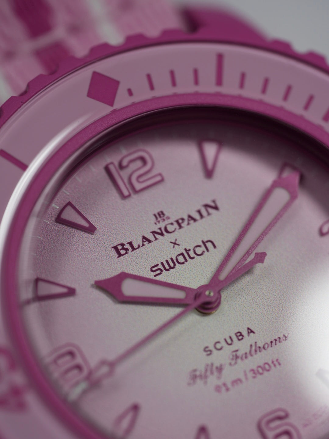 A close-up of the Blancpain x Swatch Fifty Fathoms Scuba watch in pink, highlighting the textured gradient dial with embossed numerals, the "Blancpain x Swatch" branding, and the "Fifty Fathoms Scuba" inscription. The bioceramic case and rotating bezel are also visible, with a pink and white color scheme. The hands and indices