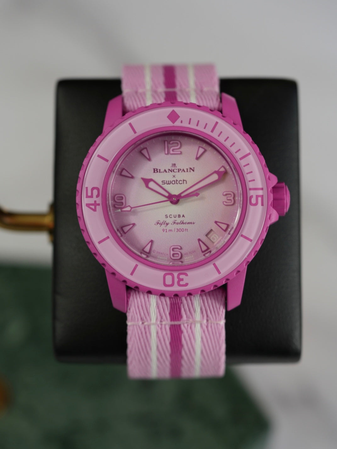 A full view of the Blancpain x Swatch Fifty Fathoms Scuba watch in pink, displayed on a black stand. The watch features a pink bioceramic case, a rotating bezel with dive markings, a gradient pink dial with embossed numerals, and a pink NATO-style strap with white stripes. The dial prominently displays the "Blancpain x Swatch" branding and "Fifty Fathoms Scuba" text, with a water resistance rating of 91m (300ft).