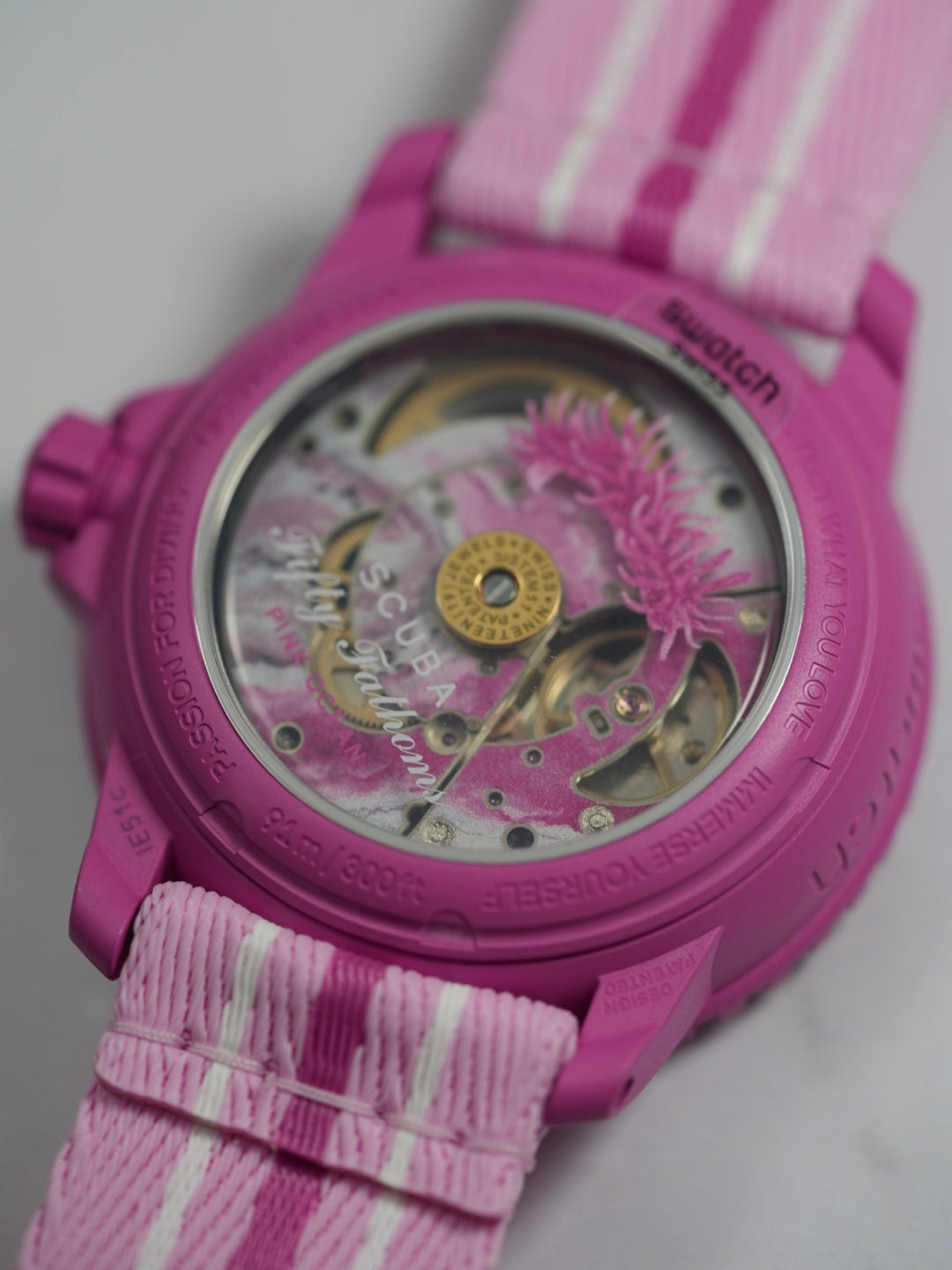 A full view of the Blancpain x Swatch Fifty Fathoms Scuba watch in pink caseback. The watch features a pink bioceramic case, a rotating bezel with dive markings, a gradient pink dial with embossed numerals, and a pink NATO-style strap with white stripes. The dial prominently displays the "Blancpain x Swatch" branding and "Fifty Fathoms Scuba" text, with a water resistance rating of 91m (300ft).