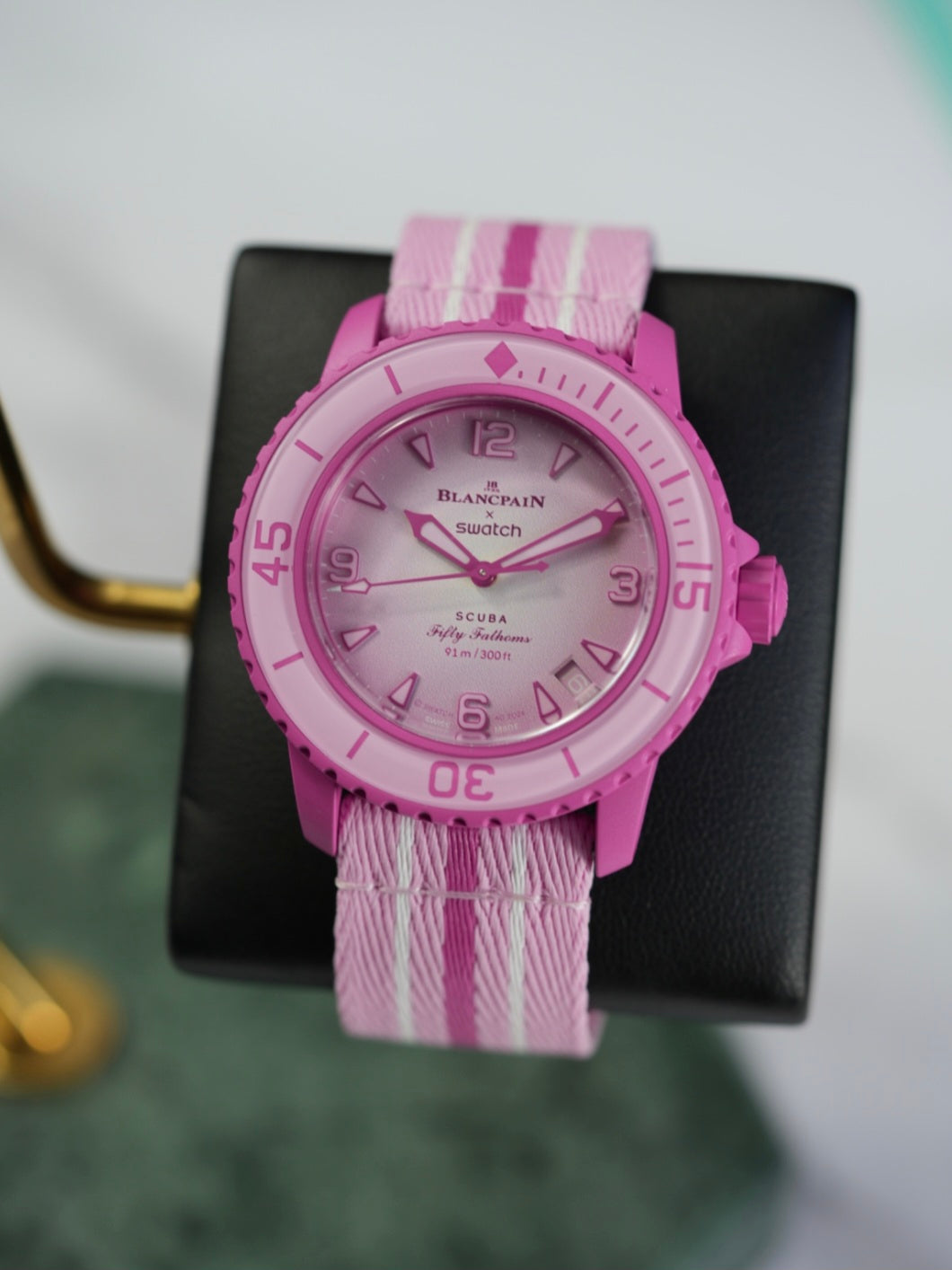 A full view of the Blancpain x Swatch Fifty Fathoms Scuba watch in pink, displayed on a black stand. The watch features a pink bioceramic case, a rotating bezel with dive markings, a gradient pink dial with embossed numerals, and a pink NATO-style strap with white stripes. The dial prominently displays the "Blancpain x Swatch" branding and "Fifty Fathoms Scuba" text, with a water resistance rating of 91m (300ft).