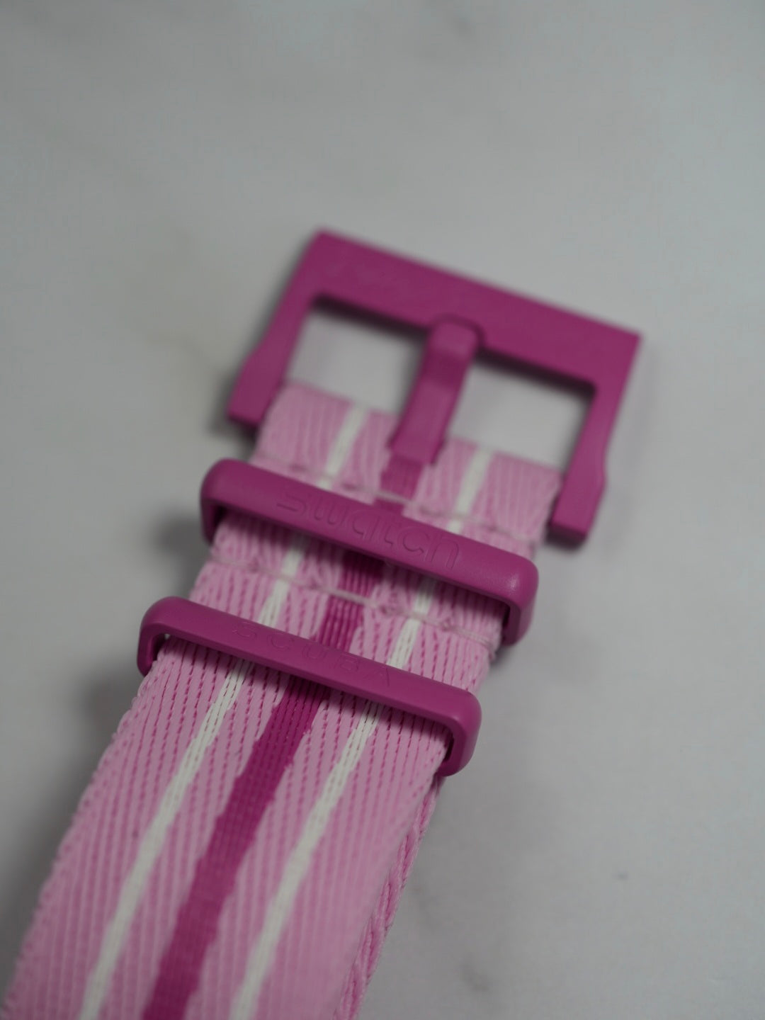 A close-up of the Blancpain x Swatch Fifty Fathoms Scuba watch strap in pink. The NATO-style fabric strap features a striped design with white and dark pink accents. The strap hardware, including the buckle and keepers, is made of pink bioceramic material, with the "Swatch" branding subtly embossed on the keepers.