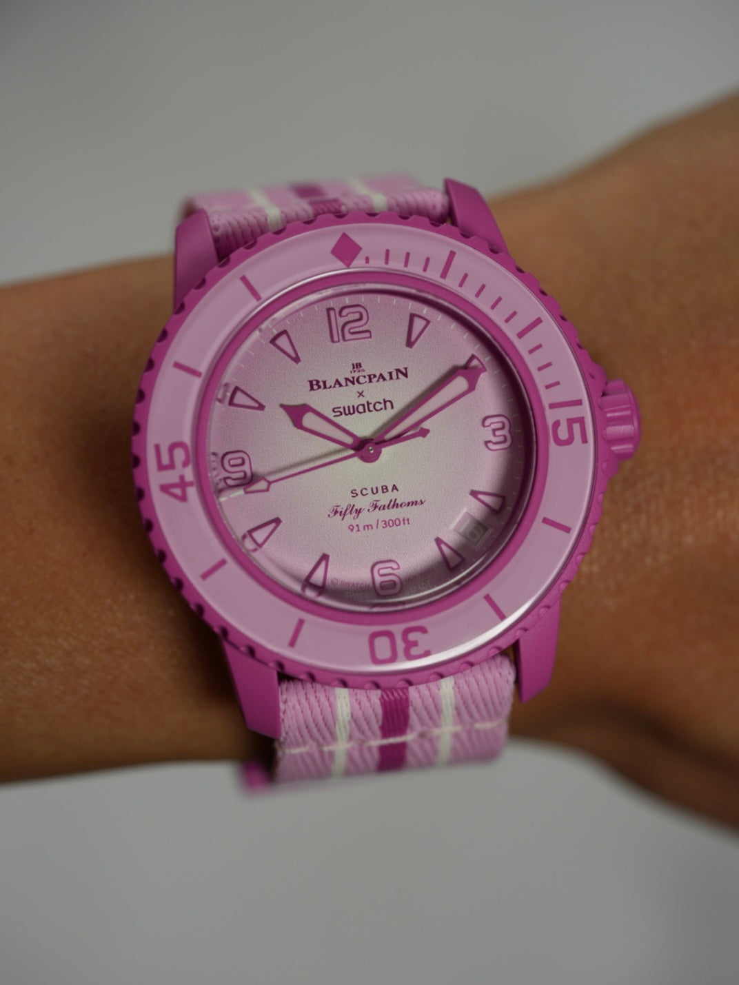 A full view on wrist the Blancpain x Swatch Fifty Fathoms Scuba watch in pink. The watch features a pink bioceramic case, a rotating bezel with dive markings, a gradient pink dial with embossed numerals, and a pink NATO-style strap with white stripes. The dial prominently displays the "Blancpain x Swatch" branding and "Fifty Fathoms Scuba" text, with a water resistance rating of 91m (300ft).