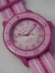 A Blancpain x Swatch Fifty Fathoms Scuba watch in vibrant pink, featuring a pink bioceramic case, a rotating bezel, a gradient pink dial with embossed numerals and indices, and a matching pink NATO-style strap with white stripes. The watch is water-resistant to 91m (300ft) and combines Swatch’s playful design with Blancpain’s iconic dive watch heritage.