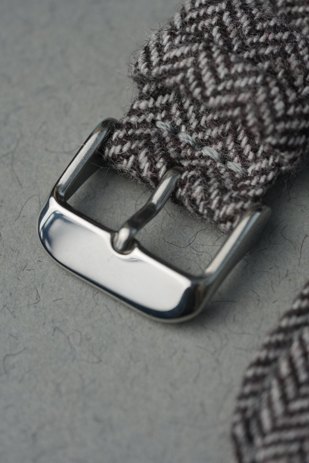 Close-up of a silver buckle on a herringbone-patterned strap against a gray background