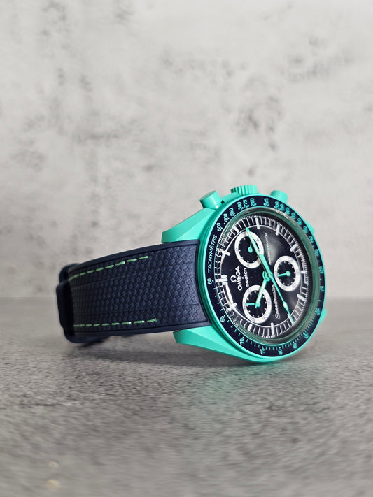 A watch with a blue case and a black woven rubber strap with green stitching on a textured background.