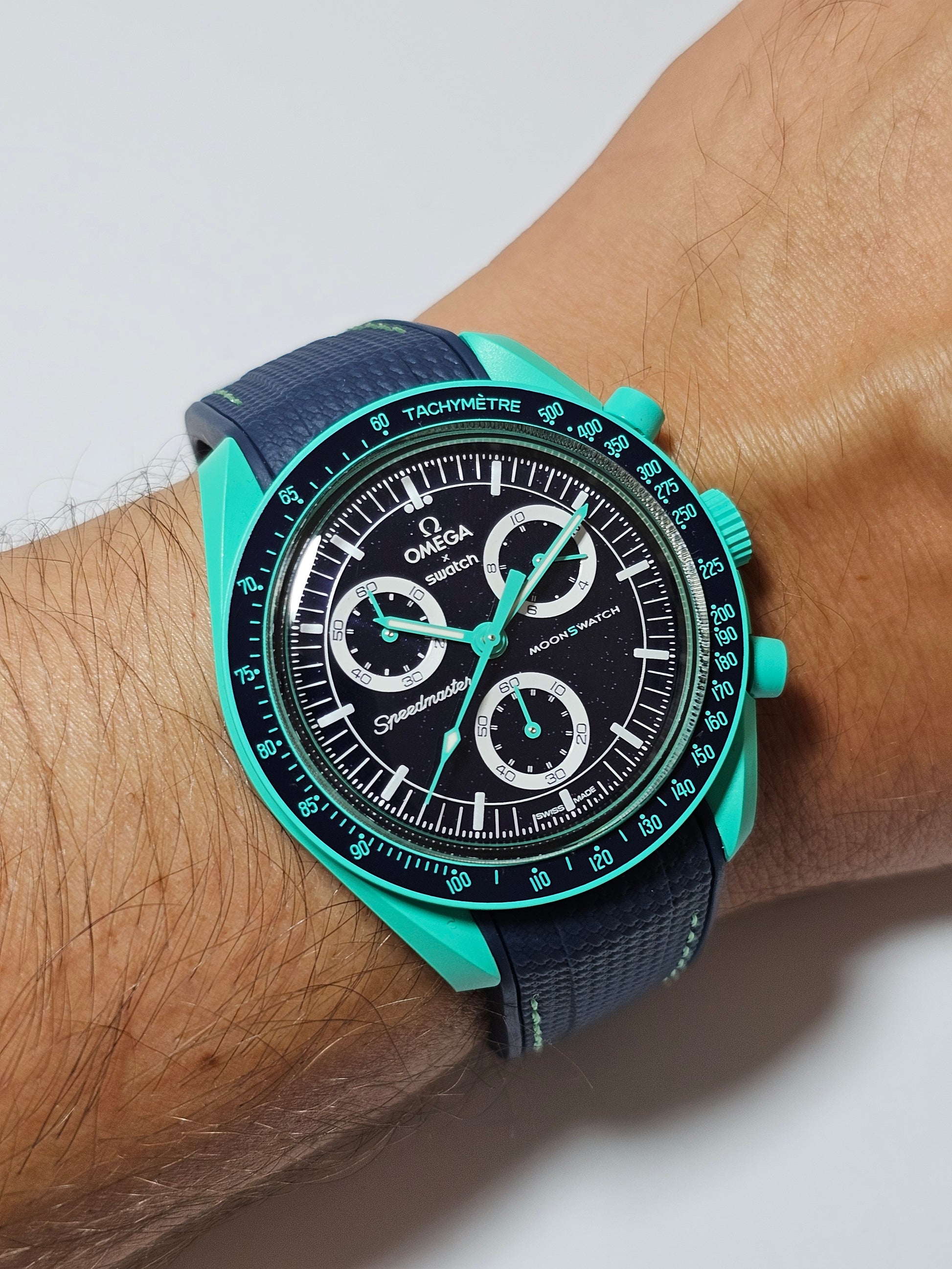 A wrist wearing a watch with a blue silicone strap and an Omega X Swatch MoonSwatch dial in polar lights mint color.