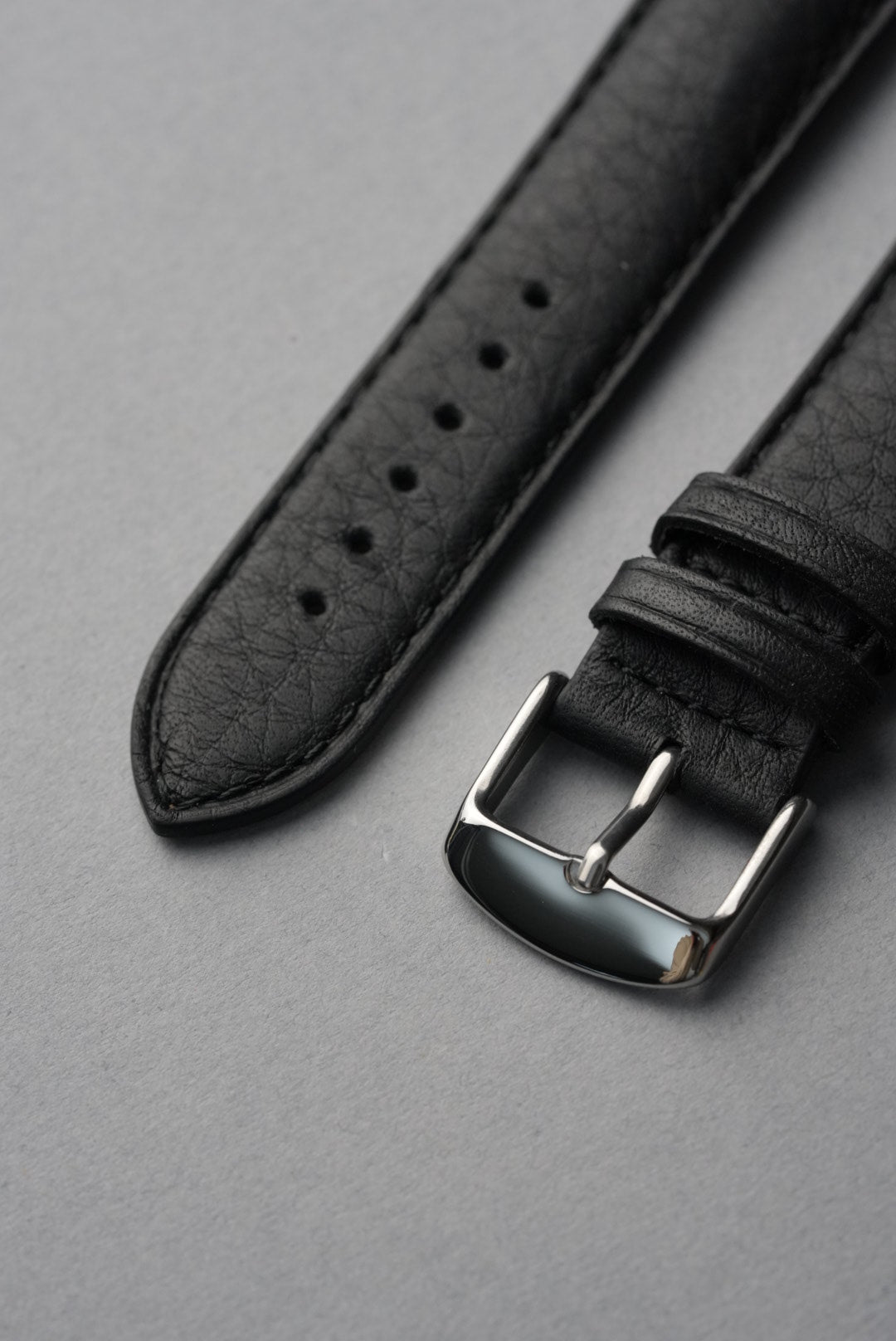 Black leather watch strap with silver buckle on a gray background