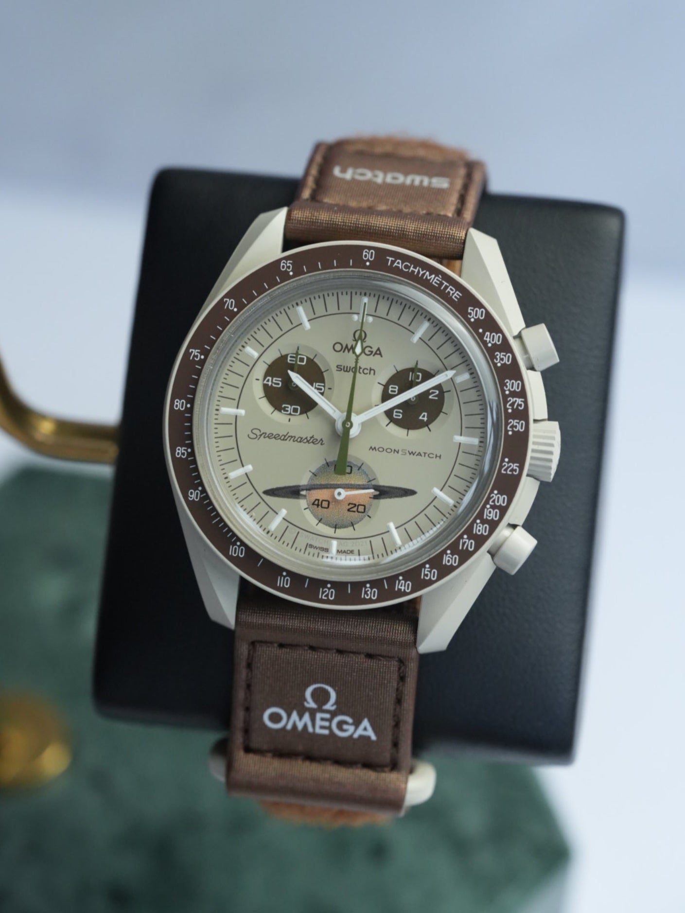 A Swatch x OMEGA MoonSwatch Collection watch with a brown leather band and a brown and silver watch face, displayed on a stand with the OMEGA logo.