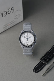 Gray 1965 MoonSwatch wristwatch with white face on a gray surface, next to a black strap and a box labeled '1965'.