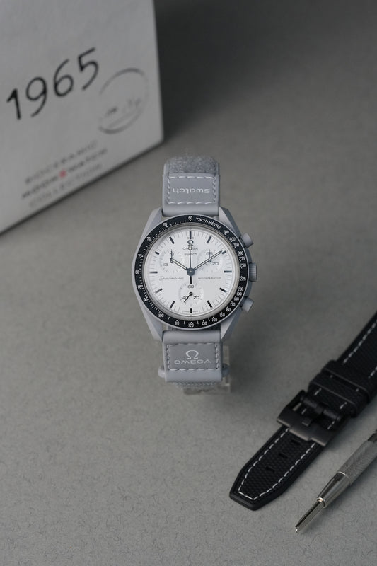 Gray 1965 MoonSwatch wristwatch with white face on a gray surface, next to a black strap and a box labeled '1965'.