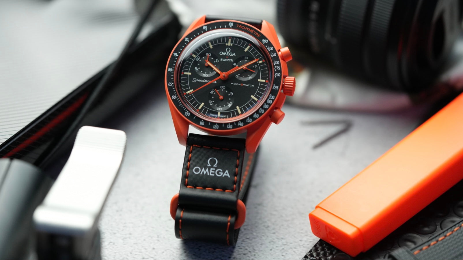 Swatch x Omega MoonSwatch Mission on Earth Lava watch with a black strap and orange stitching, displayed in a rugged setting with matching orange accessories.