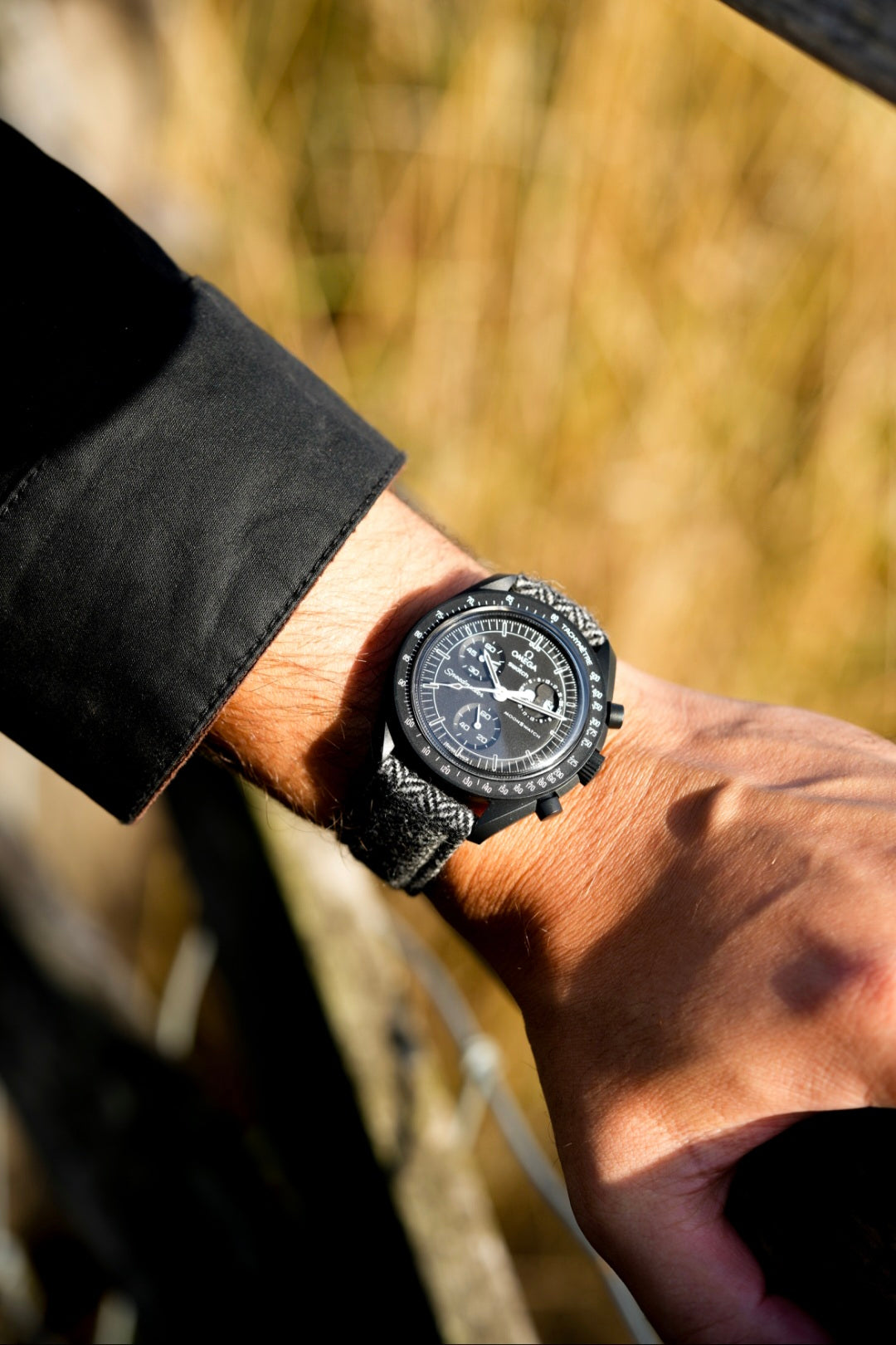 Close-up of a wrist wearing a black wristwatch with a detailed face, against a blurred natural background.