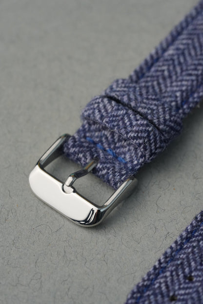 Close-up of a blue herringbone fabric strap with a silver buckle on a gray background