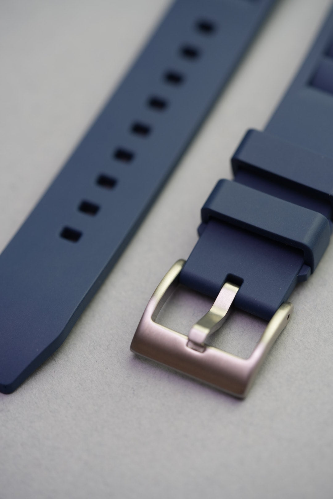 Brushed stainless steel buckle on navy vented FKM strap, shown in close-up against grey background.