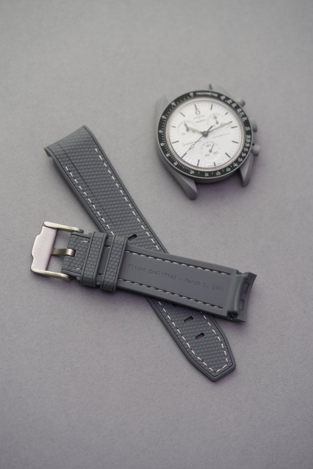 Grey Flight Qualified 1965 strap with white stitching and brushed buckle, shown next to Omega x Swatch MoonSwatch.