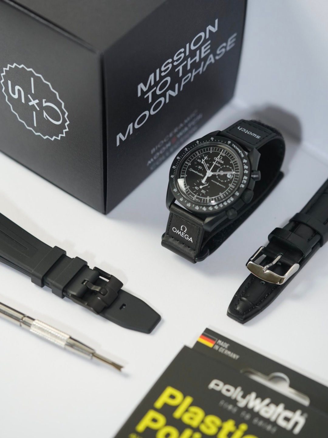 Swatch x Omega MoonSwatch Mission to the MoonPhase watch in BLACK, displayed on a stand with a matching black strap featuring Omega branding. The original packaging box labelled 'Mission to the MoonPhase' is in the background. A black textured watch strap with a stainless steel buckle, a black leather strap with white stitching, a spring bar tool, and a PolyWatch Plastic Polish kit are also arranged on a white surface.