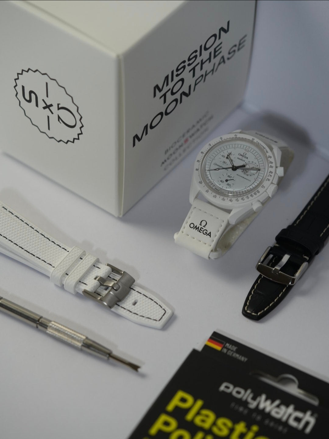 Swatch x Omega MoonSwatch Mission to the MoonPhase watch in white, displayed on a stand with a matching white strap featuring Omega branding. The original packaging box labeled 'Mission to the MoonPhase' is in the background. A white textured watch strap with a stainless steel buckle, a black leather strap with white stitching, a spring bar tool, and a PolyWatch Plastic Polish kit are also arranged on a white surface.Swatch x Omega MoonSwatch Mission to the MoonPhase watch in white, displayed on a stand wit