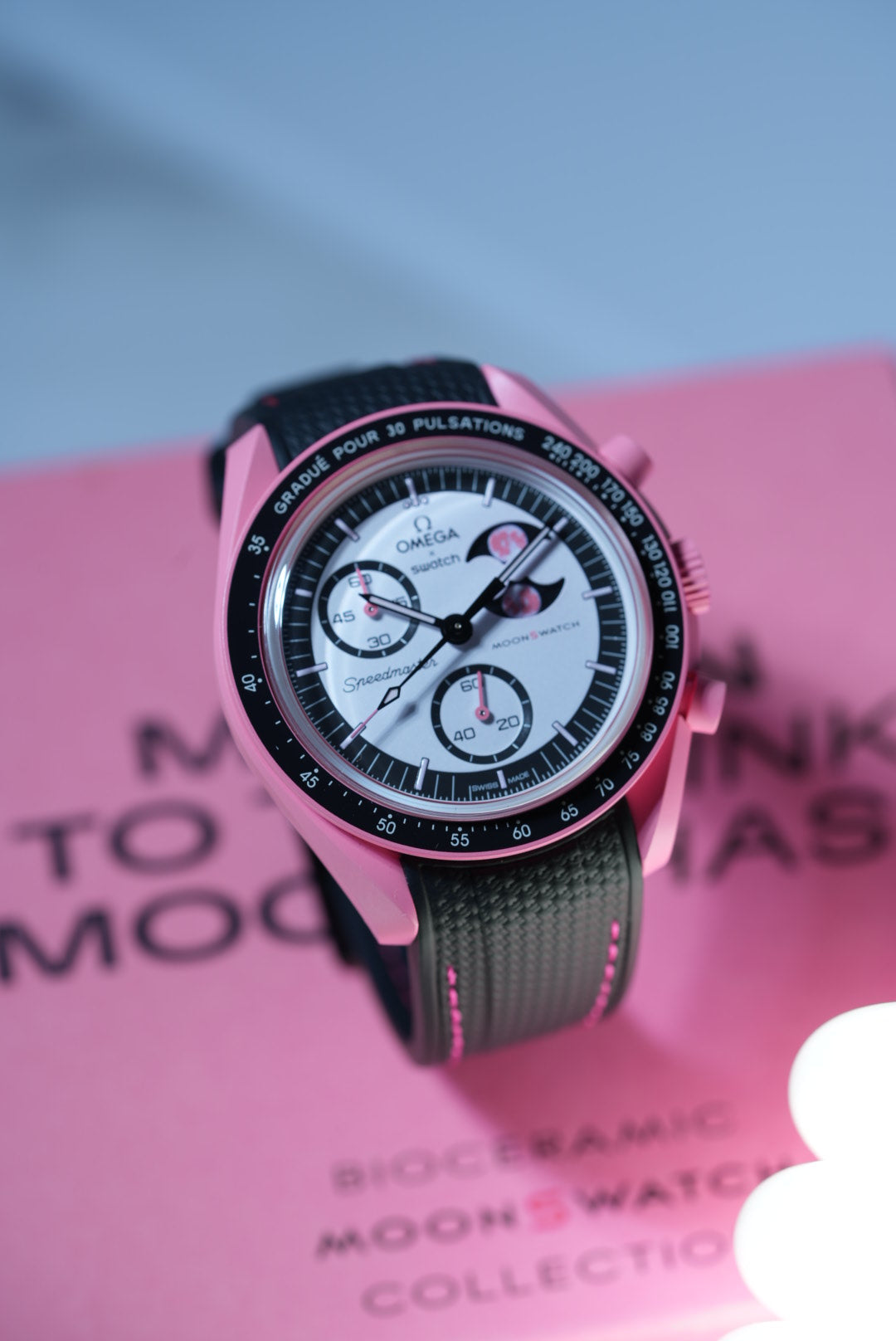 Lifestyle shot of the Pink Moonphase MoonSwatch placed in front of its original pink box – paired with a black strap and pink stitching.