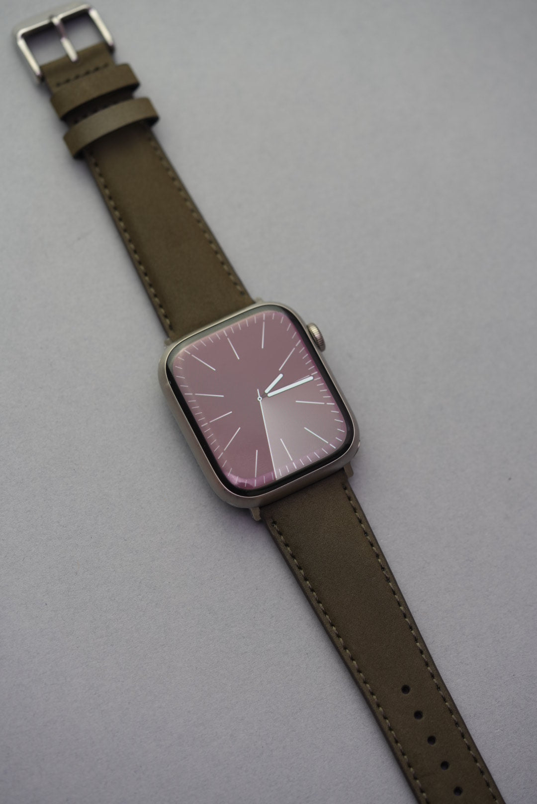Olive nubuck Apple Watch strap with polished silver buckle and visible keeper loops, shown at angle.
