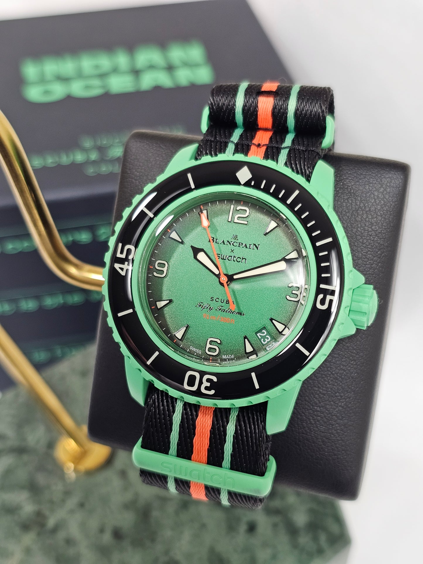 A close-up photograph of the Blancpain x Swatch Scuba Fifty Fathoms "Indian Ocean" watch displayed on a black stand. The watch features a green Bioceramic case, a black bezel with white markings, and a metallic green dial with luminescent hands and hour markers. The dial includes red text indicating "51 fathoms" as a nod to its water resistance. It is paired with a black NATO-style strap featuring green and orange stripes. In the background, a blurred box with green lettering reinforces the model's theme. T