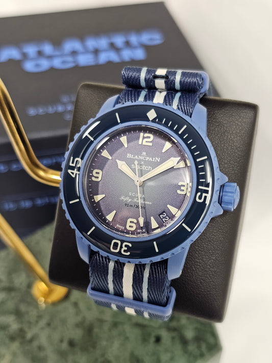 A close-up photograph of the Blancpain x Swatch Scuba Fifty Fathoms "Atlantic Ocean" watch displayed on a black stand. The watch features a deep blue Bioceramic case, a matching blue bezel with white markings, and a textured gradient blue dial with luminescent hands and hour markers. It is paired with a navy blue NATO-style strap with white and light blue stripes. In the background, a blurred box with the words "Atlantic Ocean" reinforces the model's theme. The watch is positioned on a green marble surface 