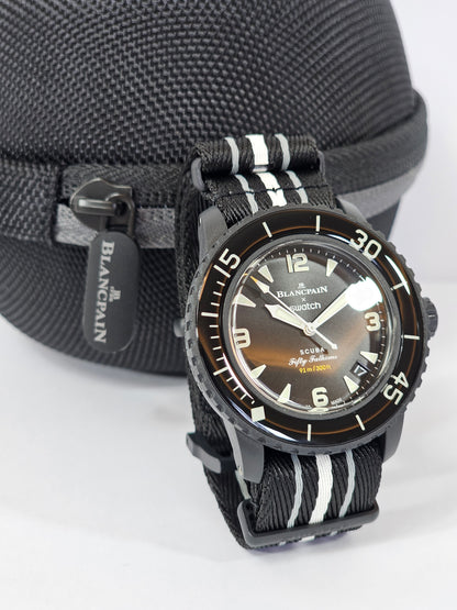  A close-up photograph of the Blancpain x Swatch Scuba Fifty Fathoms "Ocean of Storms" watch displayed upright with a black textured travel case in the background. The watch features a black Bioceramic case, a matching black bezel with white markings, and a gradient brown-to-black dial with luminescent hands and hour markers. The text on the dial includes "91m / 300ft," referencing its water resistance. It is paired with a black NATO-style strap featuring white stripes. The background includes a black trave