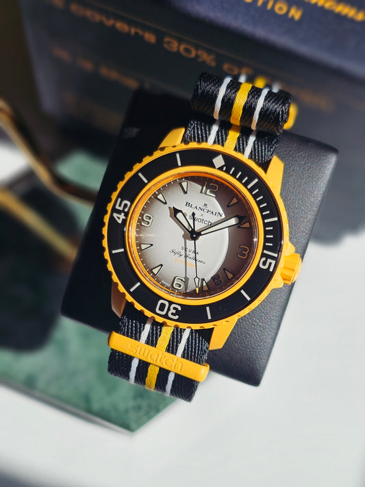 A close-up photograph of the Blancpain x Swatch Scuba Fifty Fathoms "Pacific Ocean" watch displayed on a black stand. The watch features a vibrant yellow Bioceramic case, a black bezel with white markings, and a gradient silver-to-black dial with luminescent hands and hour markers. The text on the dial includes "91m / 300ft," referencing its water resistance. It is paired with a black NATO-style strap featuring yellow and white stripes. In the background, a blurred box with gold lettering reinforces the mod