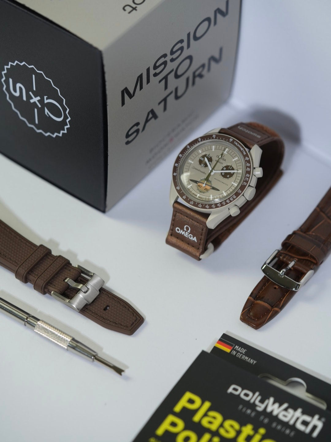 Swatch x Omega MoonSwatch Mission to Saturn watch displayed on a stand, featuring a beige bioceramic case, brown tachymeter bezel, and matching brown strap. The watch is accompanied by additional strap options, including a brown woven strap and a brown leather strap with stitching. A spring bar tool and a PolyWatch Plastic Polish kit are also included, with the original 'Mission to Saturn' packaging box in the background